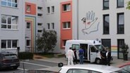 Eine Gruppe von Polizeibeamten, unter ihnen auch Mitarbeiter der Spurensicherung, steht um einen Einsatzwagen herum, der vor einem Obdachlosenheim geparkt ist. Die seitliche Schiebetür des weißen Transporters mit Blaulichtern auf dem Dach ist geöffnet, so dass Arbeitsmaterialien herausgeholt werden können. © NDR Foto: Stefan Tretropp
