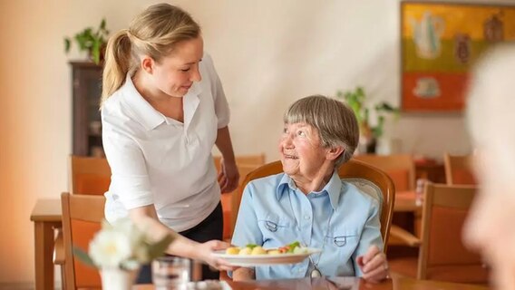 Eine Frau reicht einer Seniorin am Tisch einen Teller zum Mittagessen. © Kursana 