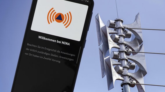 Nahaufnahme einer Sirene an einer Schule, daneben die geöffnete Warn-App NINA auf einem Smartphone © Udo Herrmann 