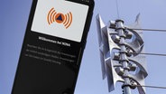 Nahaufnahme einer Sirene an einer Schule, daneben die geöffnete Warn-App NINA auf einem Smartphone © Udo Herrmann 