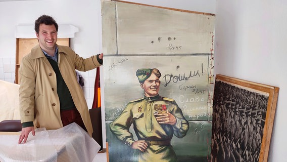 Jakob Schwichtenberg ist Historiker und steht nebem einem Gemälde, auf dem ein Sowjetsoldat zu sehen ist, dazu Worte in kyrillischen Buchstaben. © NDR Foto: Martin Möller