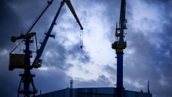 Dunkle Wolken ziehen über das Gelände der MV Werften mit Krananlagen, einem ehemaligen Wohnschiff für Werftarbeiter und der großen Schiffbauhalle.  