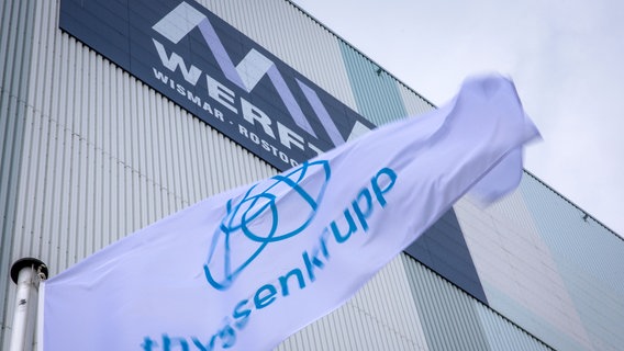 Die Fahne des Unternehmens ThyssenKrupp weht vor der Schiffbauhalle auf dem Gelände der Werft in Wismar, an der noch der Schriftzug MV Werften zu sehen ist. © dpa Bildfunk Foto: Jens Büttner