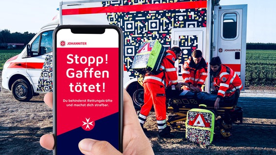 Ein Rettungswagen ist mit einem übergroßen QR-Code beklebt. Davor hält eine Hand ein Smartphone, auf dem "Stopp! Gaffen tötet!" steht. © Johanniter-Unfall-Hilfe 