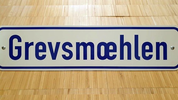 Plattdeutsches Ortsschild für Grevesmühlen: Auf weißem Grund steht in blauen Buchstaben "Grevsmœhlen". © NDR Foto: Thomas Naedler