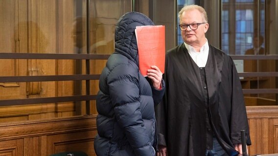Der Angeklagte hält sich im Gerichtssaal eine Mappe vor das Gesicht. Neben ihm steht sein Verteidiger. © Paul Zinken/dpa Foto: Paul Zinken/dpa