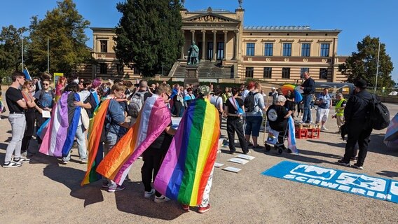 Zahlreiche, vor allem junge Leute stehen bei einer Demonstration gegen Queerfeindlichkeit in bunte Fahnen gehüllt auf dem Alten Garten in Schwerin. © NDR Foto: Christoph Loose