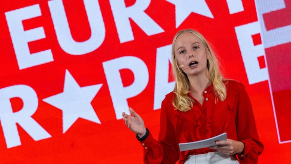 Die 24-jährige Sabrina Repp stellt sich vor ihrer Wahl zur Kandidatin aus Mecklenburg-Vorpommern mit einer Bewerbungsrede vor. Die blonde junge Frau steht in einer roten Bluse vor einem rot-weißen Europawahl-Banner. © dpa Foto: Jens Büttner