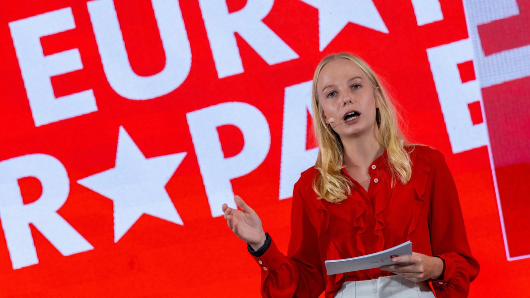 Die 24-jährige Sabrina Repp stellt sich vor ihrer Wahl zur Kandidatin aus Mecklenburg-Vorpommern mit einer Bewerbungsrede vor. Die blonde junge Frau steht in einer roten Bluse vor einem rot-weißen Europawahl-Banner. 