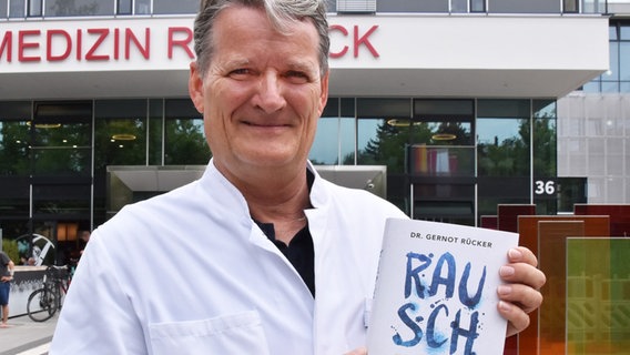 Ein Mann steht vor einem Klinikgebäude und hält ein Buch mit dem Titel "Rausch" in die Kamera. © Unimedizin Rostock Foto: Unimedizin Rostock