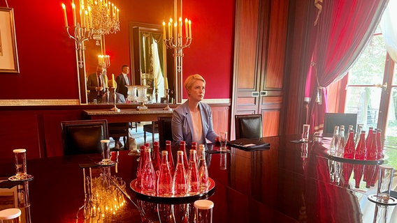 Schwesig in der Residenz des deutschen Botschafters in Paris. Sie sitzt an einem großen Tisch in einem Zimmer mit dunkelroten Wänden. Durch ein bodentiefes Fenster dringt Tageslicht an schweren roten Vorhängen vorbei in den Raum. Auf dem Tisch stehen Wasserflaschen und Gläser, hinter Schwesig hängen Kerzenständer und ein Spiegel, in dem man ihre Begleiter sieht, die noch nicht sitzen. Neben sich auf dem Tisch hat Schwesig eine Mappe liegen. © NDR MV Foto: Anna-Lou Beckmann