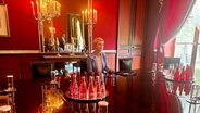 Schwesig in der Residenz des deutschen Botschafters in Paris. Sie sitzt an einem großen Tisch in einem Zimmer mit dunkelroten Wänden. Durch ein bodentiefes Fenster dringt Tageslicht an schweren roten Vorhängen vorbei in den Raum. Auf dem Tisch stehen Wasserflaschen und Gläser, hinter Schwesig hängen Kerzenständer und ein Spiegel, in dem man ihre Begleiter sieht, die noch nicht sitzen. Neben sich auf dem Tisch hat Schwesig eine Mappe liegen. © NDR MV Foto: Anna-Lou Beckmann