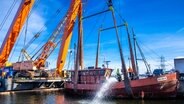 Ein riesiger orangener Kran hebt ein tonnenschweres Schiff aus dem Rostocker Hafenbecken. Gleichzeitig stehen Arbeiter in Warnwesten auf dem Deck des Schiffs und pumpen Wasser aus dem Rumpf. Alles passiert unter einem blauen Himmel. © dpa/Jens Büttner Foto: Jens Büttner