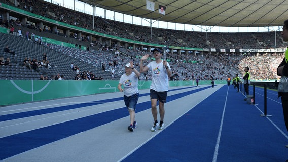 Zwei Menschen, die Special Olympics T-Shirts tragen, laufen in Berlin beim DFB-Pokalfinale auf der Laufbahn des Stadions. © IMAGO images Foto: IMAGO / Schüler