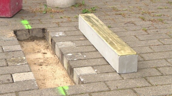 Eine Stolperschwelle liegt vor einem vorbereitetem Loch zwischen Pflastersteinen im Boden.  