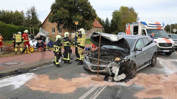 Feuerwehrmänner und weitere Einsatzkräfte stehen vor einem demolierten Unfallwagen vor dem eine Ölspur mit Sand abgedeckt wurde. © NDR Foto: Stefan Tretropp