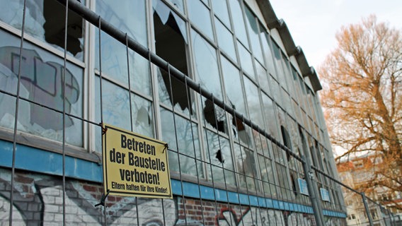 Hinter einem Gitterzaun mit der Aufschrift "Betreten der Baustelle verboten" steht die verfallende Meerwasserschwimmhalle mit eingeworfenen Scheiben. © NDR.de Foto: Daniel Sprenger