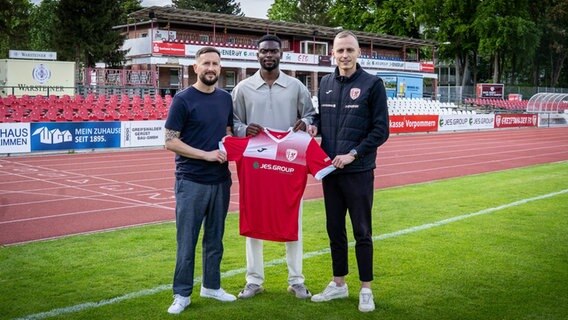 Greifswalder FC feiert Neuzugang von Moordy Hüther. Er hält Trikot mit Trainer und Geschäftsstellenleiter. © Greifswalder FC Foto: Greifswalder FC