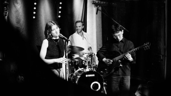 Eine Frau steht vor einem Mikrofon und singt. Daneben ein Mann mit einer Gitarre in der Hand. Im Hintergrund ist ein weiterer Mann an einem Schlagzeug zu sehen. © Žan Vidmar Zorc 