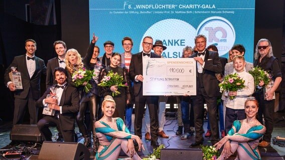 Ein Gruppe Menschen steht auf einer Bühne und hält einen Spenden-Scheck. © Windflüchter Gala 