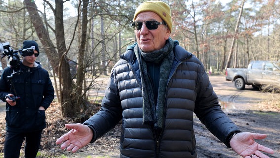 Rüdiger Voßhall, Gründer der Regenbogen AG, spricht bei einem Pressetermin auf dem Campingplatz. Er trägt eine Steppjacke, eine gelbe Mütze und eine Sonnenbrille und hält die Arme in einer Geste scheinbarer Ahnungslosigkeit von sich. © dpa Foto: Bernd Wüstneck