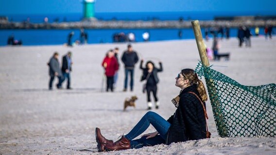 eine junge Frau sitzt im Sand an einem Zaunpfahl am Strand von Warnemünde. © dpa Foto: Jens Büttner