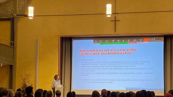 Etwa 50 Menschen sitzen und schauen auf eine Leinwand. Vorne steht eine Frau, die einen Vortrag hält. © Jakob Gaberle 