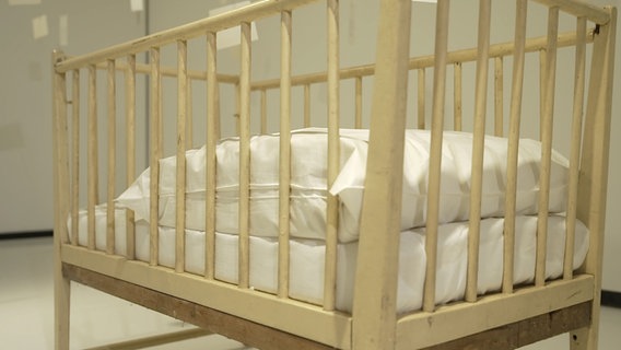 Das Bild zeigt ein Kinderbett in einer Ausstellung der Kunsthalle Rostock. © NDR Foto: Screenshot