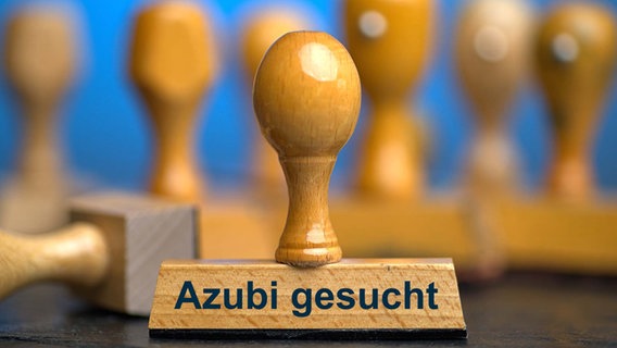 Ein Holzstempel mit der Aufschrift "Azubi gesucht" auf einer Schieferplatte mit weiteren Stempeln unscharf dahinter. © picture alliance/Torsten Sukrow/SULUPRESS.DE 