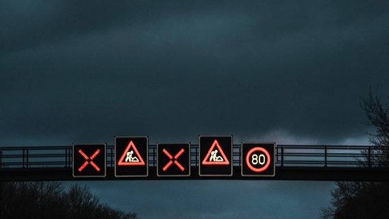 Eine bei Dunkelheit beleuchtete Schilderbrücke mit Baustellenschildern und Tempolimitanzeige über einer Autobahn. © NDR Foto: Julius Matuschik