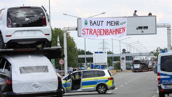 Aktivisten von verschiedenen Klimaschutzgruppen befestigen ein Transparent mit der Aufschrift "Baut mehr Straßenbahnen" an einer Schilderbrücke über der Liefereinfahrt am Stammwerk von Volkswagen. © dpa Foto: Bodo Marks