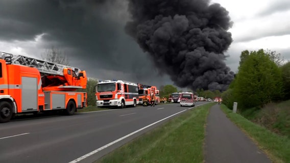 In einer Straße stehen viele Einsatzfahrzeuge der Feuerwehr und im Hintergrund steigt eine Rauchsäule auf. © aktuell24 