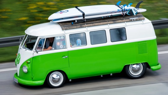 Auf einer Straße fährt ein alter VW-Bulli mit zwei Surfbrettern auf dem Dach. © dpa - Bildfunk Foto: Peter Steffen