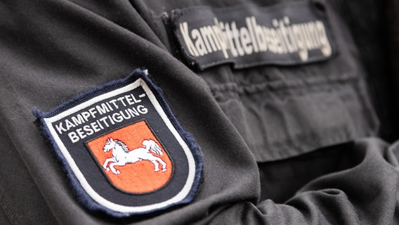 Ein Sprengmeister trägt Dienstkleidung mit der Aufschrift "Kampfmittelbeseitigung" und dem Niedersachsen-Ross. © picture alliance/dpa Foto: Swen Pförtner