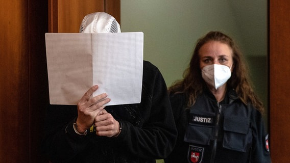 Der Angeklagte kommt mit einem Schreibblock vor dem Gesicht und Handschellen zur Urteilsverkündung im Mordprozess ins Landgericht. © Swen Pförtner/dpa Foto: Swen Pförtner