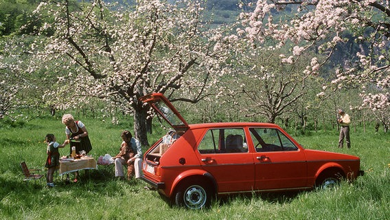 Ein roter VW-Golf steht inmitten von blühenden Bäumen und eine Familie macht ein Picknick. © dpa - Bildarchiv 