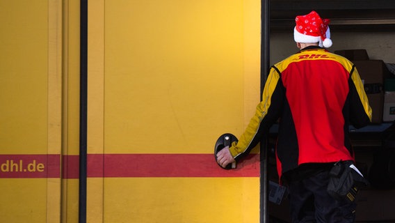 Vor einem Lieferwagen von DHL steht ein Paketzusteller mit einer Weihnachtsmütze auf dem Kopf. © dpa - Bildfunk Foto: Julian Stratenschulte