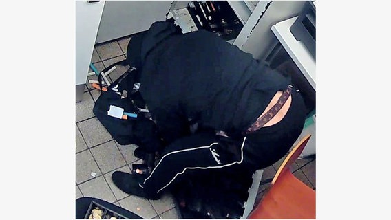 Ein in schwarz gekleideter Räuber steht gebückt über einer Tasche, daneben liegt Bargeld. Ein in schwarz gekleideter Räuber steht gebückt über einer Tasche, daneben liegt Bargeld. © Polizei Göttingen