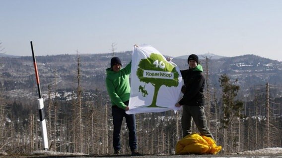 Zwei Menschen halten ein Banner mit der Aufschrift "40 - Robin Wood", im Hintergund sind abgestorbene Bäume zu sehen. © Robin Wood Foto: Moritz Heck