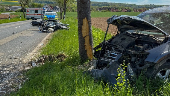 im Vordergrund steht ein stark beschädigtes Auto auf einem Grünstreifen neben einem Baum. Auf der Straße liegen ein zerstörtes Motorrad und ein kaputtes Leichtkraftrad, dahinter sind ein Rettungswagen und ein Streifenwagen zu sehen. © Konstantin Mennecke/Kreisfeuerwehr Northeim Foto: Konstantin Mennecke