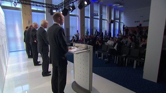 Vorstände von Volkswagen stehen auf einer Bühne an Rednerpulten. © NDR 