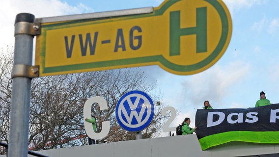 Mehrere Greenpeace-Aktivisten stehen auf dem Eingang der VW-Zentrale in Wolfsburg. Im Vordergrund ist ein Haltestellenschild mit der Aufschrift "VW-AG" zu sehen. © dpa - Bildfunk Foto: Peter Steffen