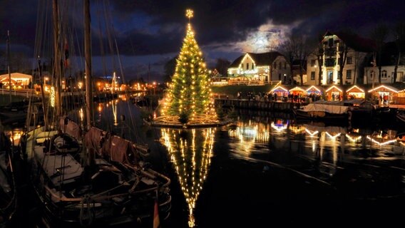 Schwimmender Weihnachtsbaum im Hafen von Carolinensiel bei Mondschein. Rechts Beleuchtung von Weihnachtsmarktständen. © NDR Foto: Marion Holtz