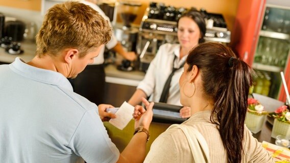 Ein Mann und eine Frau stehen in einem Cafe am Tresen und schauen auf eine Rechnung. © MODEL RELEASED, MR, PROPERTY RELEASED, PR Foto: MODEL RELEASED, MR, PROPERTY RELEASED, PR
