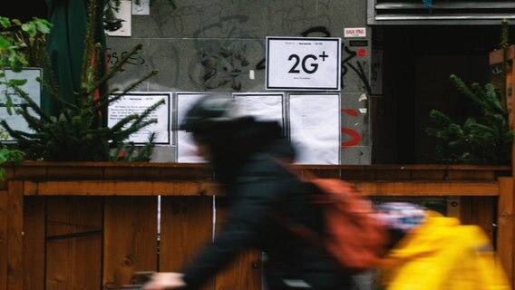 Das Bild zeigt einen Radfahrer der an einem Ladengeschäft vorbeifährt. Im Schaufenster hängt ein Schild, dass auf die 2G-Plus Regelung hinweist. © picture alliance / NurPhoto | Ying Tang Foto: Ying Tang