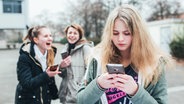 Ein Mädchen schaut auf ihr Handy, im Hintergrund lachen zwei weitere Mädchen. © klicksafe.de Foto: Maribelle Photography