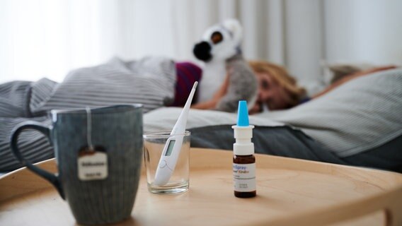 Fieberthermometer, Nasenspray und eine Tasse Tee stehen vor einem Bett, in dem ein Kind mit Kuscheltier liegt. © dpa Foto: Annette Riedl