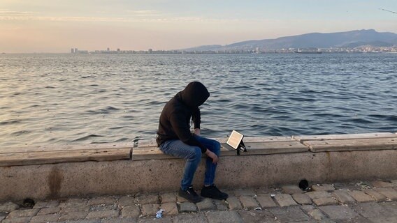 Eine Person mit Kapuze sitzt auf einer Mauer vor einem Gewässer. © NDR Foto: Angelika Henkel