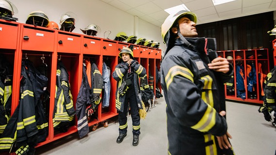 Zwei Feuerwehrmänner ziehen sich in einem Umkleideraum ihre Einsatzkleidung an. © NDR Foto: Julius Matuschik
