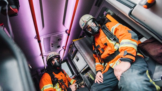Zwei Feuerwehrleute mit Atemschutzmasken sitzen in einem Einsatzfahrzeug. © NDR Foto: Julius Matuschik
